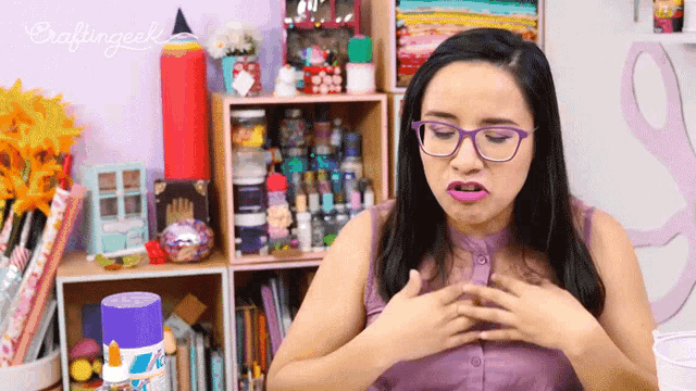 a woman wearing glasses holds her hands to her chest in front of a crafting shelf