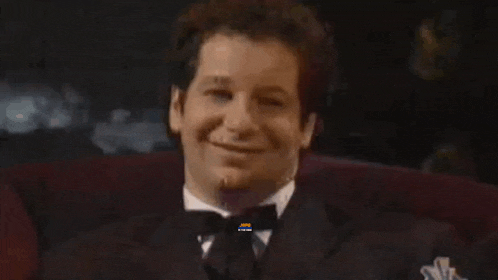 a man in a tuxedo and bow tie is smiling while sitting in a chair .