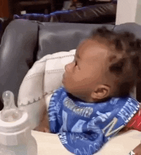 a baby wearing a bib is sitting in a high chair next to a bottle of milk .