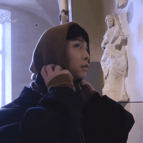 a person wearing a hooded jacket stands in front of a statue with the word paris on the wall