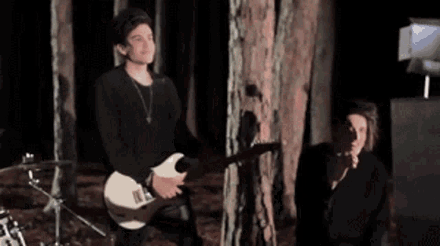 a man is playing a guitar in the woods while a woman sits in the shadows .