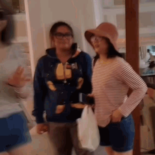 two women are standing next to each other in a room . one of the women is wearing a hat .