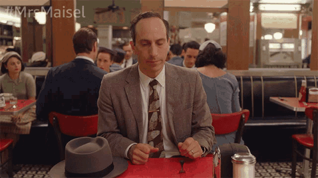 a man in a suit and tie sits at a table in a diner with the hashtag #mr maisel