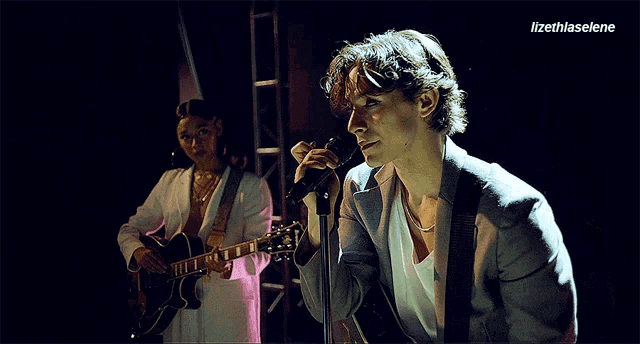 a man singing into a microphone while a woman plays a guitar in the background