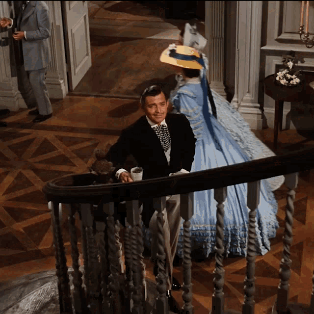 a man in a suit stands on a staircase next to a woman in a blue dress and hat
