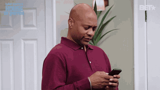 a man in a maroon shirt is looking at his phone with a house of payne logo behind him