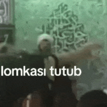 a man in a white hat is standing in front of a green wall with arabic writing .