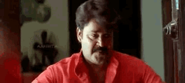 a man with a mustache is wearing a red shirt and looking at the camera .