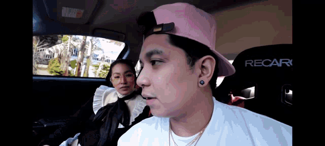 a man wearing a pink hat sits in a car next to a woman wearing a recar seat