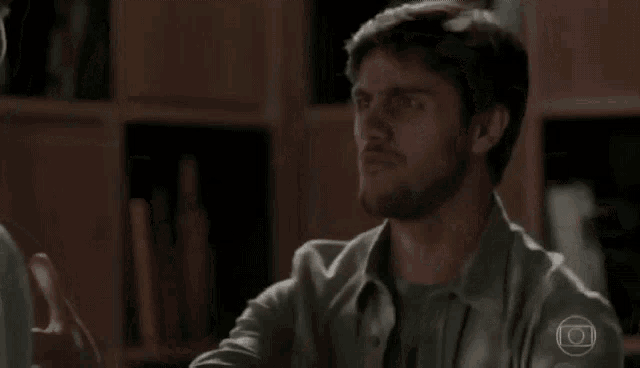 a man with a beard is sitting in front of a bookshelf and talking to another man .