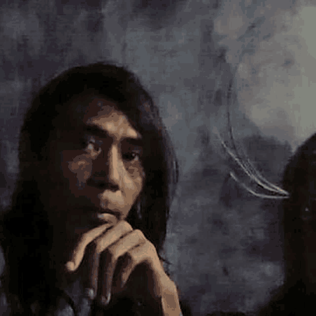 a man with long hair is sitting down with his hands on his chin .