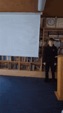 a man standing in front of a projector screen with a clock on the wall behind him
