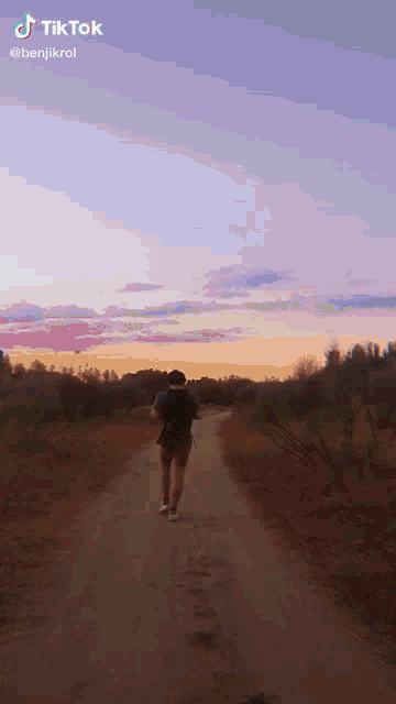 a man is walking down a dirt road with a sunset in the background