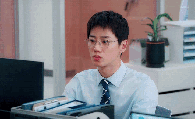 a man wearing glasses and a tie sits at a desk in front of a computer