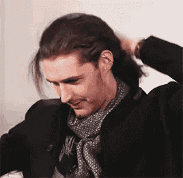a man with long hair is wearing a scarf and a jacket and holding his hair in a ponytail .