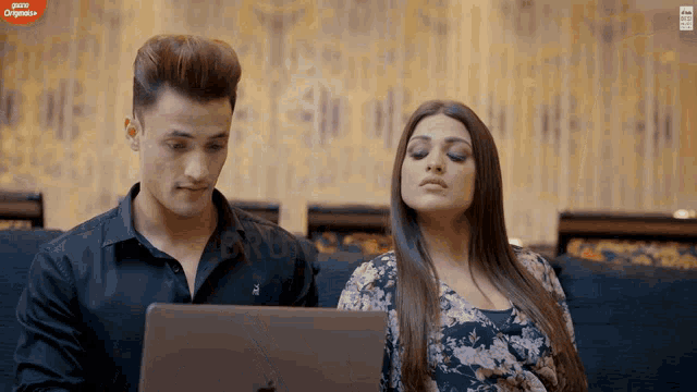 a man and a woman sit on a couch looking at a laptop screen that says ' gusto originals ' on it