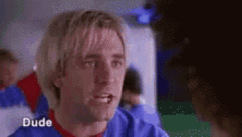 a man with blonde hair and a blue shirt is talking to another man in a room .