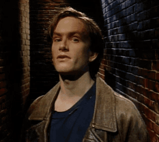 a man wearing a brown leather jacket and a blue shirt is standing next to a brick wall