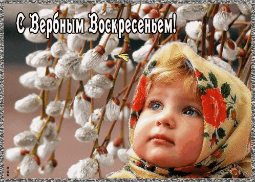 a little girl in a scarf stands in front of a bunch of flowers with a greeting in russian