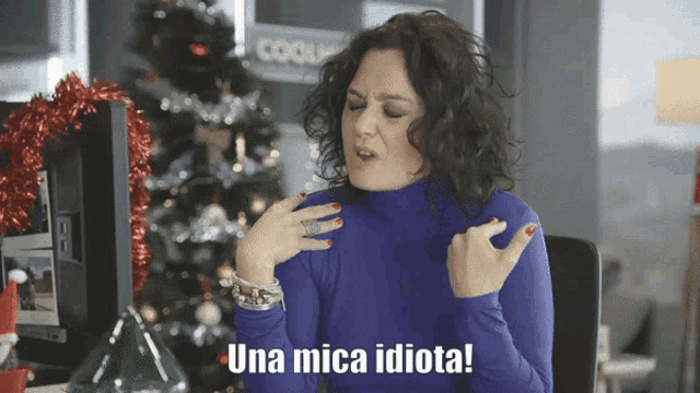 a woman in a blue turtleneck says una mica idiota in front of a christmas tree