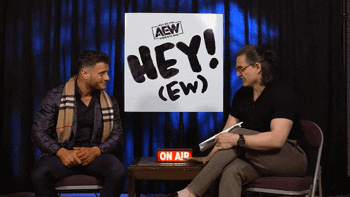 two men sitting in front of a sign that says hey