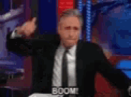 a man in a suit and tie is standing in front of a microphone with his arms outstretched and says boom .