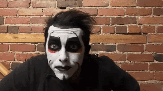 a man with black and white face paint is standing in front of a red brick wall