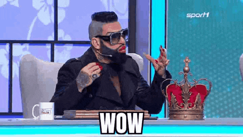 a man with a beard and red lipstick is sitting at a table with a crown .
