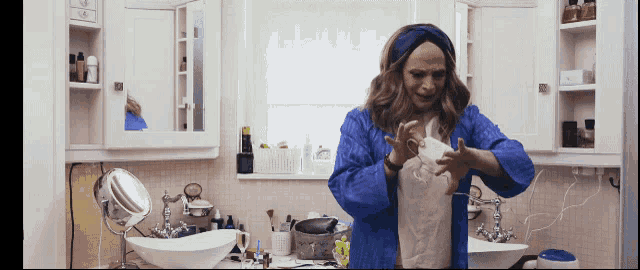 a woman in a blue robe is standing in a bathroom looking at herself in the mirror