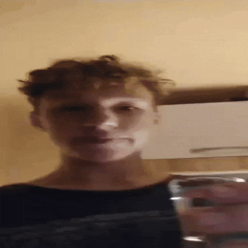 a young man with curly hair holds a glass of water in front of his face