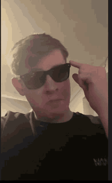 a man wearing sunglasses and a black shirt is pointing his finger at his forehead .