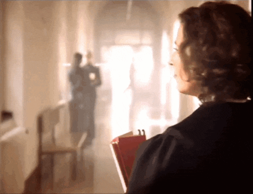 a woman holding a book in a hallway with a man in the background