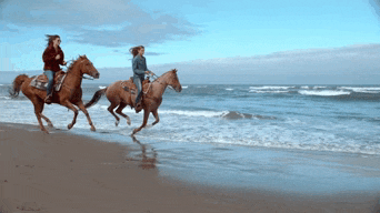 two women are riding horses on the beach near the ocean