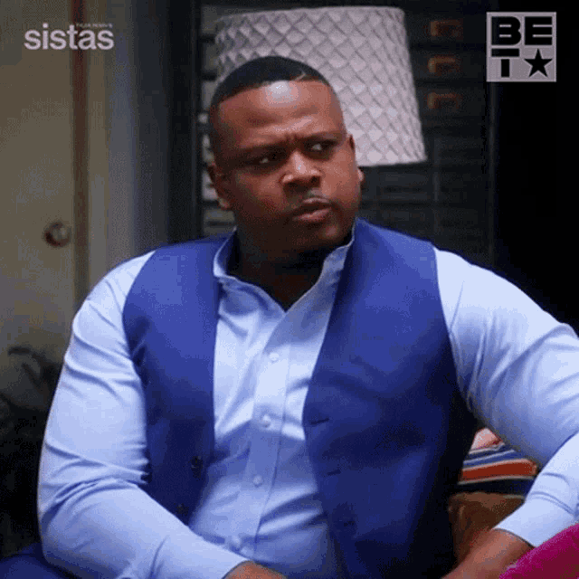 a man wearing a blue vest and a blue shirt is sitting in front of a sign that says sistas