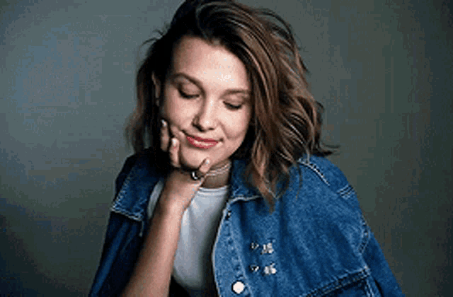 a woman wearing a denim jacket and a white shirt is smiling with her hand on her chin .
