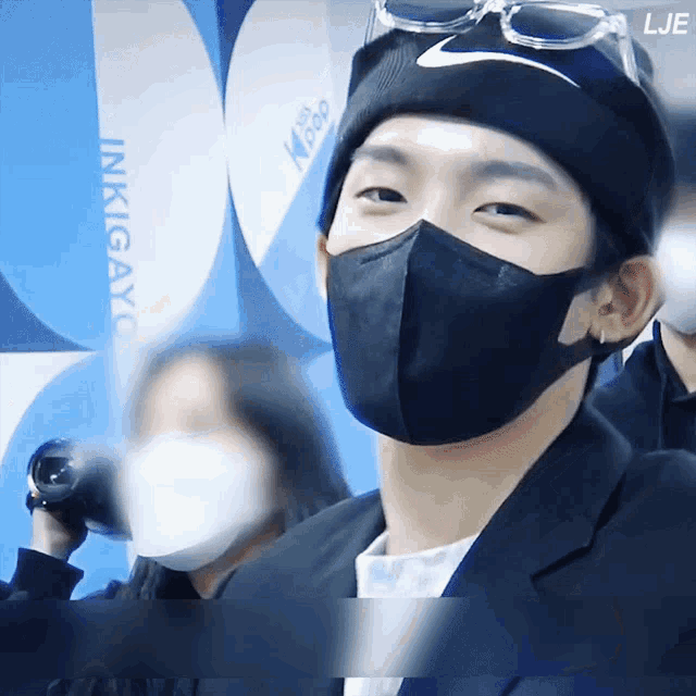 a man wearing a black face mask and a nike hat stands in front of a sign that says inkigayo