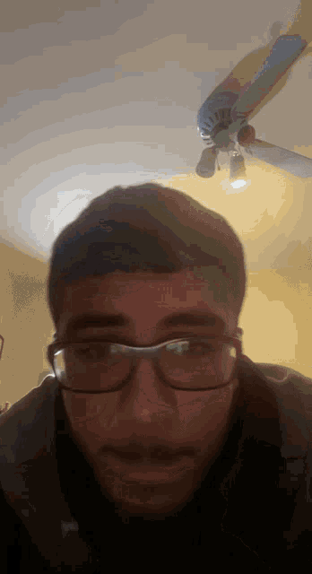a man wearing glasses looks at the camera with a ceiling fan behind him