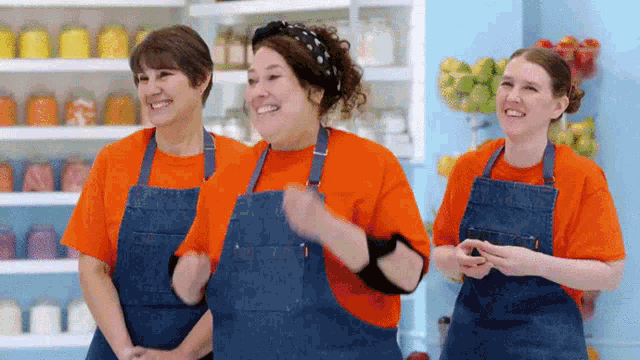 three women in orange shirts and blue aprons are standing next to each other and smiling .