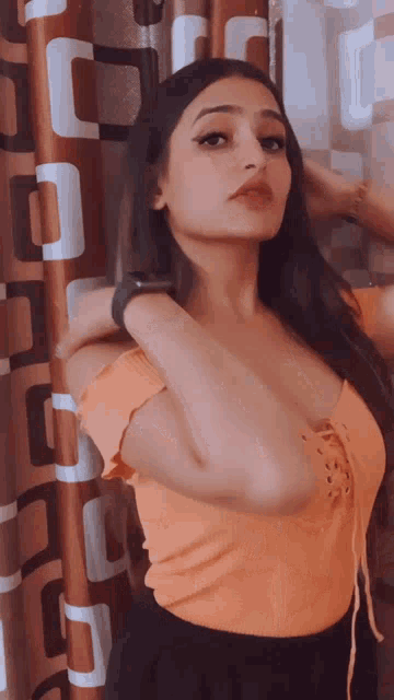 a woman wearing an orange top is standing in front of a patterned curtain
