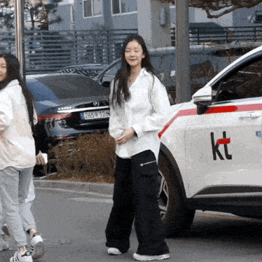 a woman in a white shirt walks past a car that says kt