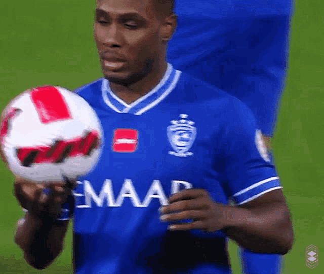 a man in a blue emaar jersey holds a soccer ball in his hand