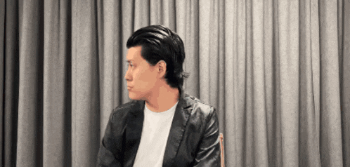 a man in a black leather jacket is sitting in front of a curtain .