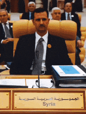 a man in a suit and tie sits in front of a microphone in front of a sign that says syria on it
