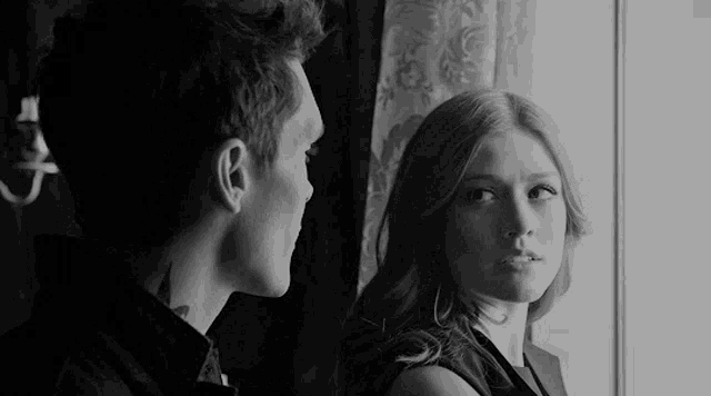 a black and white photo of a man and a woman looking at each other in front of a window .