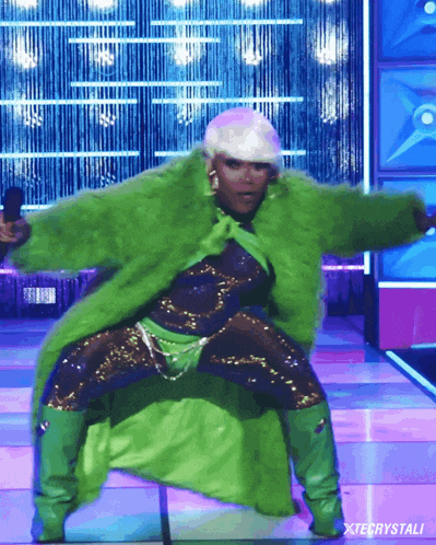 a woman in a green fur coat is dancing on a stage with xtecrystali written below her