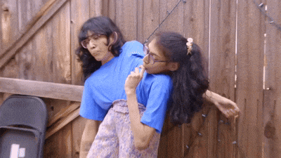 a woman in a blue shirt holds another woman on her shoulders in front of a wooden fence