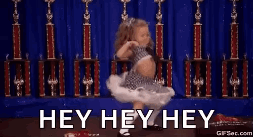 a little girl is dancing in front of trophies with the words `` hey hey hey '' written above her .