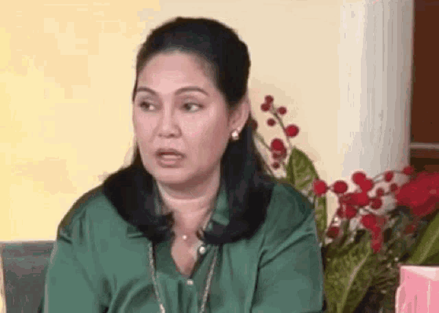 a woman in a green shirt and earrings is sitting in front of a vase of flowers .