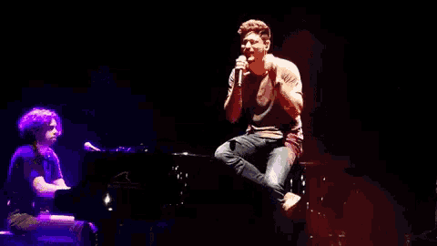 a man is sitting on a stage singing into a microphone while a man plays a piano in the background .