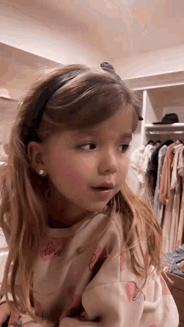 a little girl wearing a headband and earrings is standing in a closet looking at something .